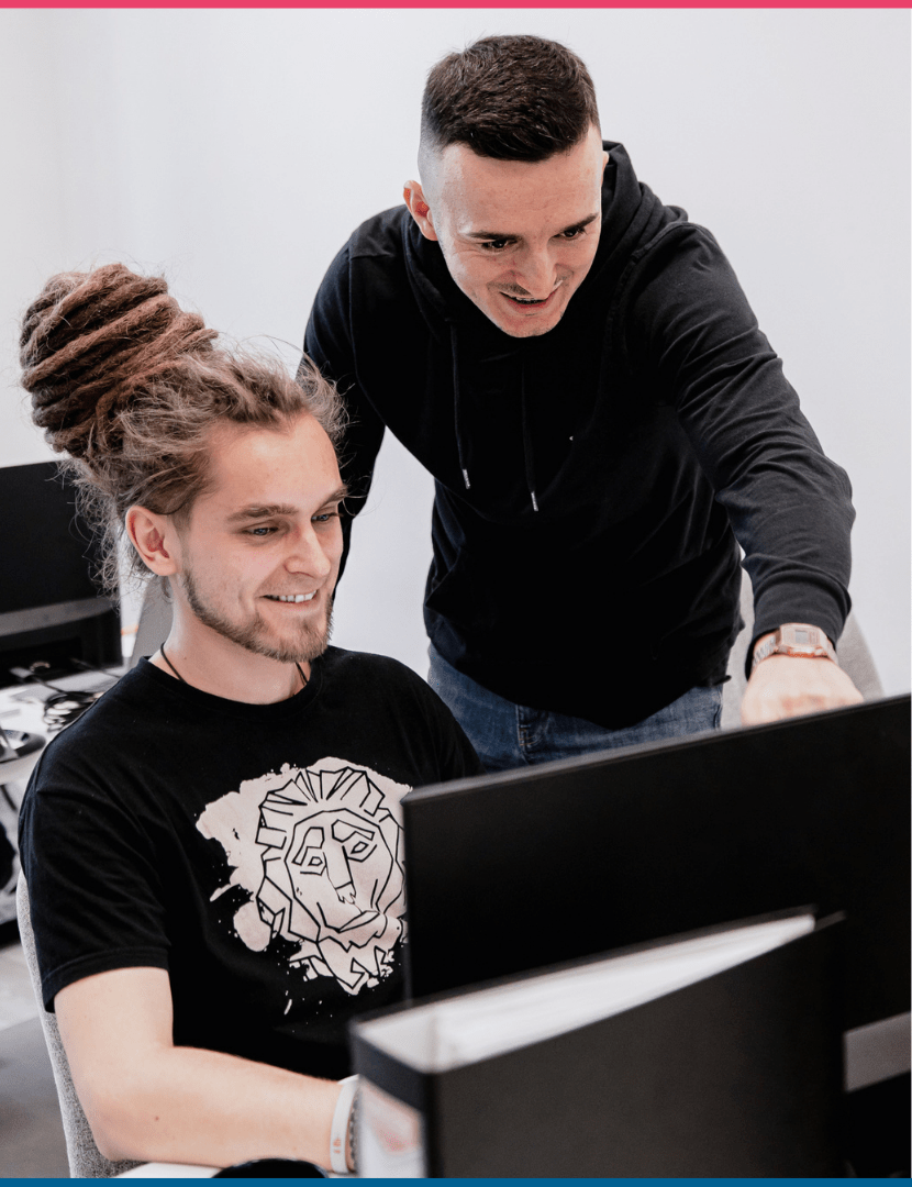 delegowanie zadań pracownikom - młody mężczyzna wskazuje palcem coś w komputerze drugiemu młodemu mężczyźnie