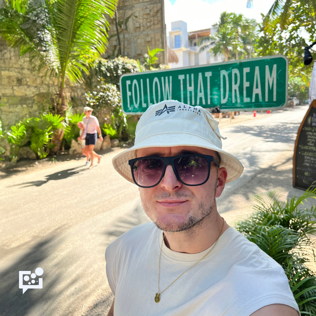 selfie młodego chłopaka w podróży, w okularach i kapeluszu na głowie, w tle napis: follow that dream